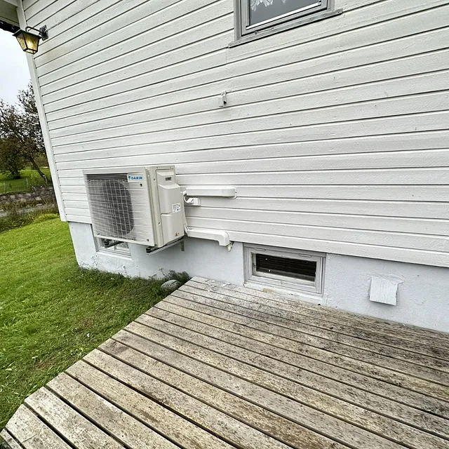 Varmepumpe utedel på lys husvegg med treplatting og grønn plen