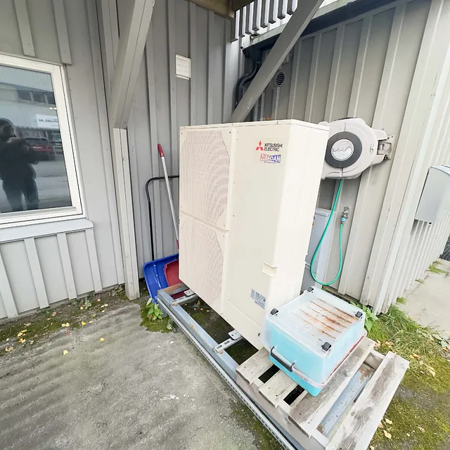 Varmepumpe utedel montert på grå husvegg ved inngang