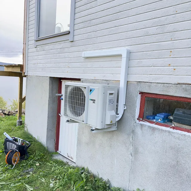 Varmepumpe utedel montert på lys husvegg med gress og små busker foran
